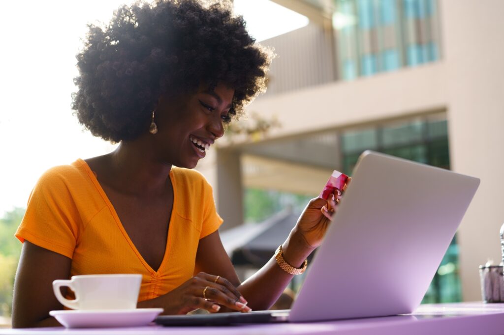 Happy young african woman holding credit card while shopping via laptop sitting in outdoor cafe