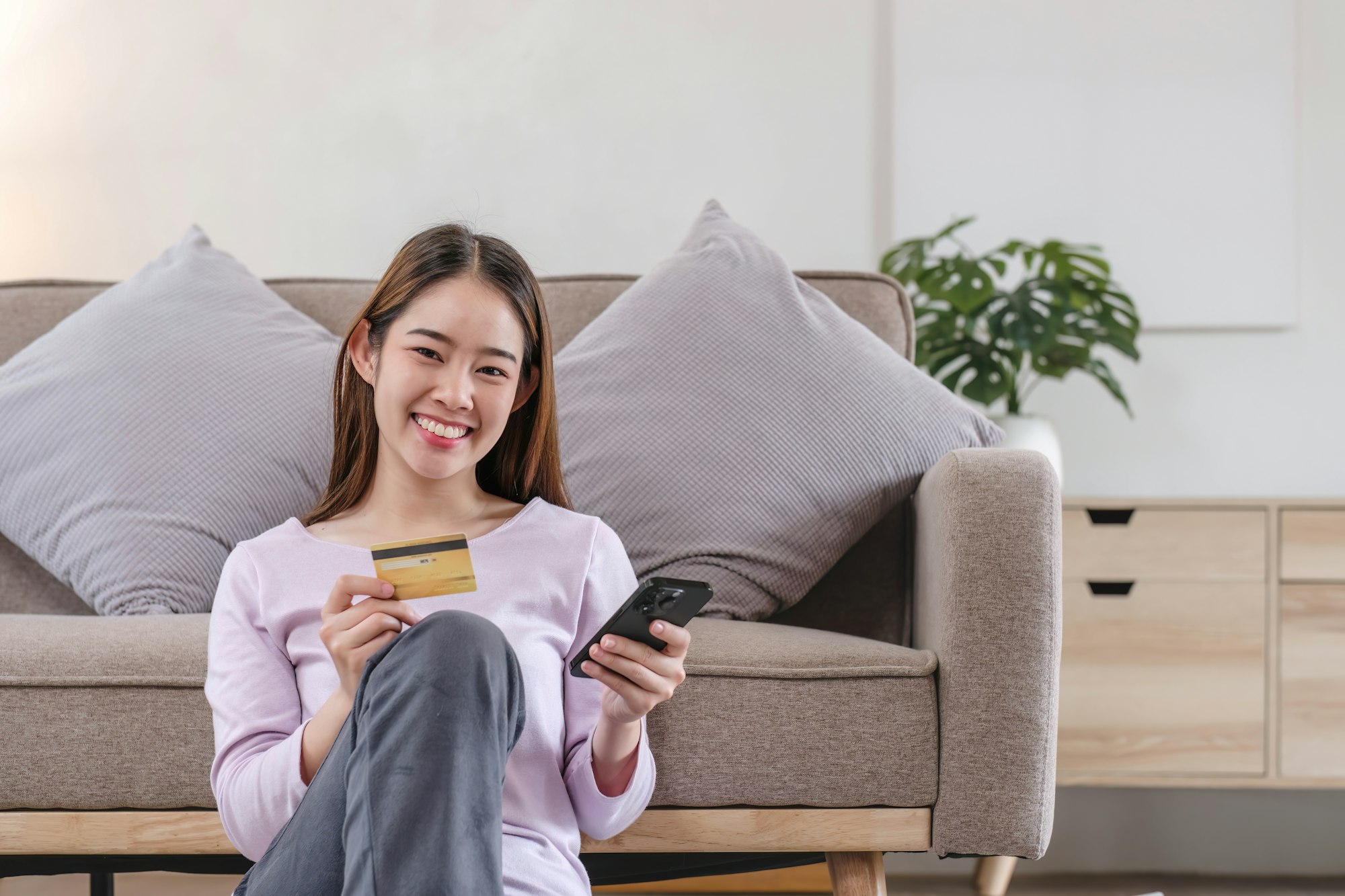 Young adult asian female consumer holding credit card and smartphone sitting on floor at home doing